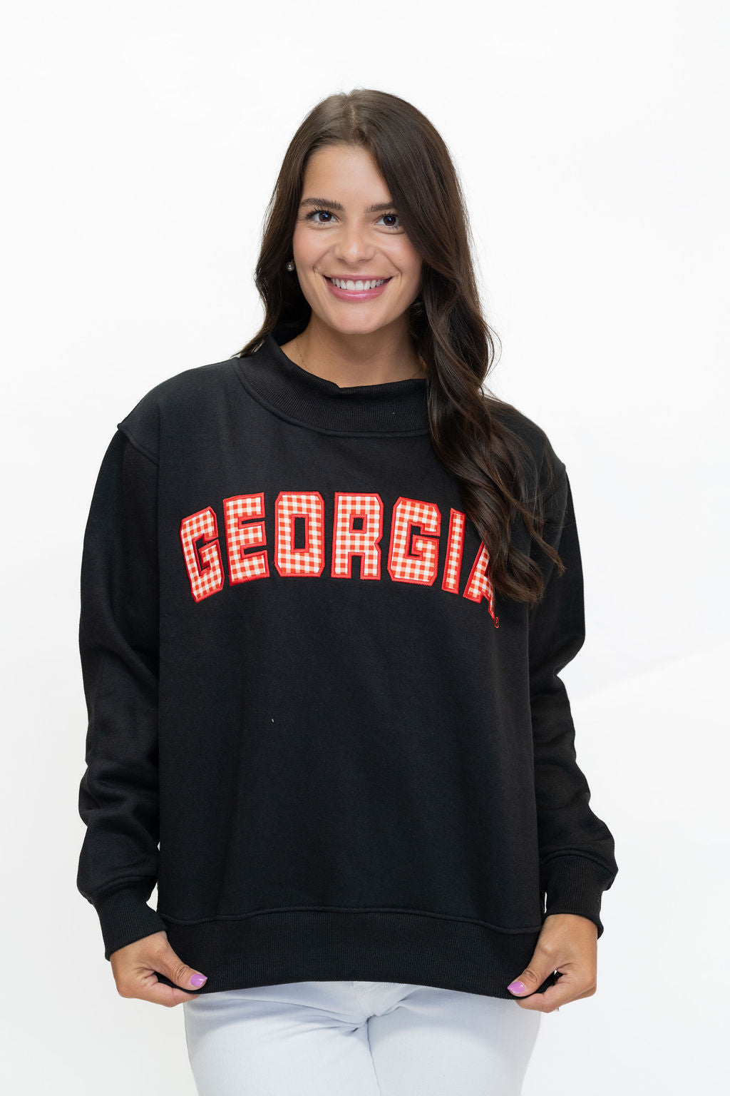 A woman wearing a black crewneck sweatshirt with 'GEORGIA' printed across the chest in red and white gingham block letters, paired with white pants, against a plain white background.