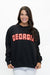 A woman wearing a black crewneck sweatshirt with 'GEORGIA' printed across the chest in red and white gingham block letters, paired with white pants, against a plain white background.
