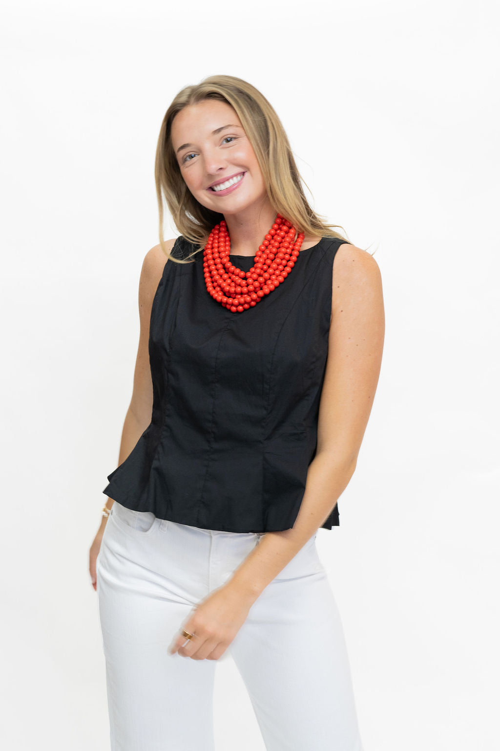 A smiling woman wearing a sleeveless black peplum top and white pants, accessorized with a chunky multi-strand red beaded necklace, photographed against a plain white studio background.