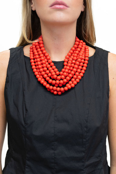 A close-up of a model wearing a multi-strand coral red beaded necklace over a black sleeveless top, set against a plain white background.