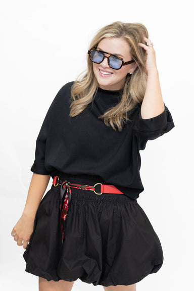 A woman modeling a black short-sleeve mock neck top and matching black pleated mini skirt, styled with a red patterned belt and black sunglasses, against a plain white background.