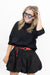 A woman modeling a black short-sleeve mock neck top and matching black pleated mini skirt, styled with a red patterned belt and black sunglasses, against a plain white background.