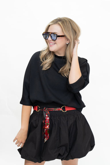 A woman modeling a black long-sleeve top and black balloon shorts with a red patterned belt, wearing tortoiseshell sunglasses against a plain white background.