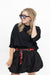 A woman modeling a black long-sleeve top and black balloon shorts with a red patterned belt, wearing tortoiseshell sunglasses against a plain white background.