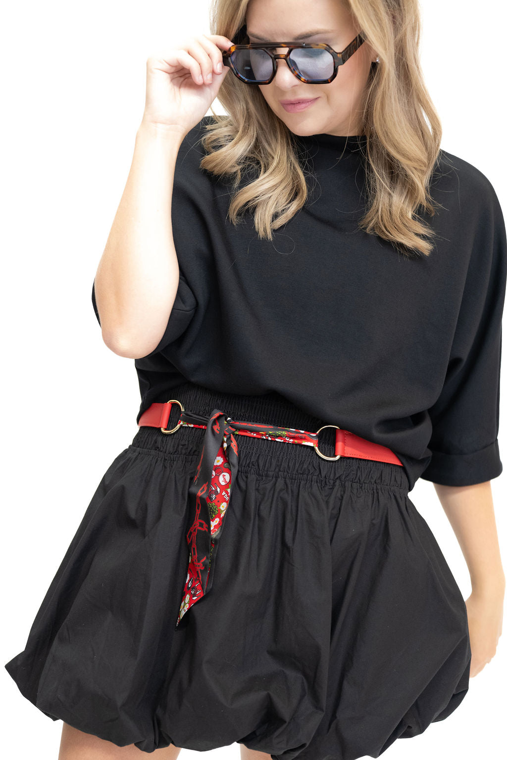 A woman wearing a black short-sleeve top and black balloon-style shorts, styled with a red belt featuring a red, white, and black patterned scarf, and black sunglasses, against a plain white background.