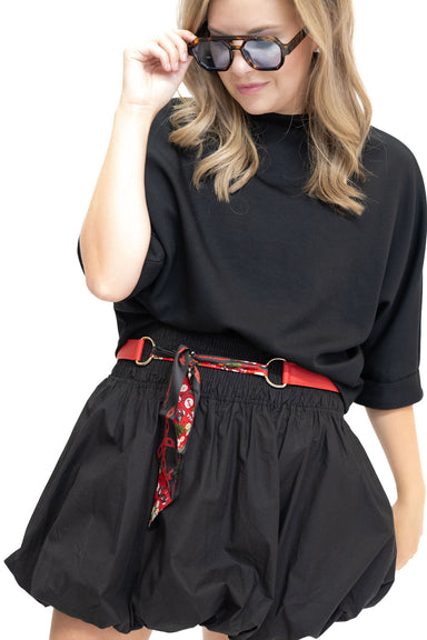 A woman wearing a black short-sleeve top and black balloon-style shorts, styled with a red belt featuring a red, white, and black patterned scarf, and black sunglasses, against a plain white background.