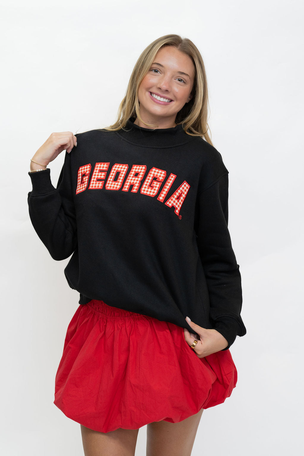 A woman modeling a black long-sleeve sweatshirt with 'GEORGIA' in red block letters outlined in white across the chest, paired with a red mini skirt, against a plain white background.