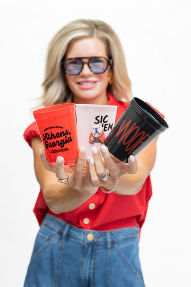A woman in a red shirt and blue jeans holds three custom-printed cups toward the camera against a white background. The cups are red, white, and black, featuring the text 'Sic Em' and 'Others, Georgia'.