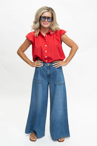 A woman modeling a bright red short-sleeve button-up shirt with ruffled shoulders and blue wide-leg denim jeans, standing against a plain white studio background.