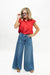 A woman modeling a red short-sleeve button-up shirt and high-waisted, wide-leg blue denim jeans against a plain white studio background.