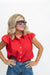 A woman with blonde hair wearing a bright red short-sleeve button-up shirt and blue jeans, posing with her hands on her hips against a plain white background.