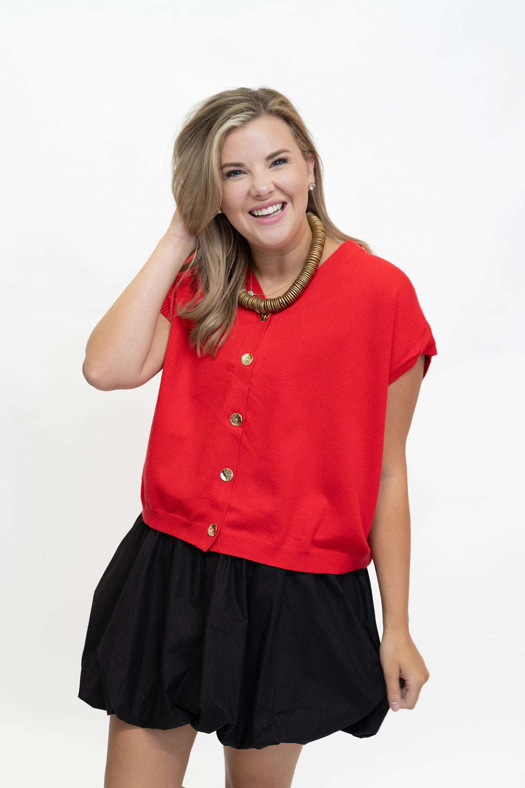 A woman modeling a bright red short-sleeve top with gold buttons down the front, paired with a black pleated skirt, against a plain white studio background.