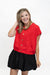 A woman modeling a bright red short-sleeve top with gold buttons down the front, paired with a black pleated skirt, against a plain white studio background.