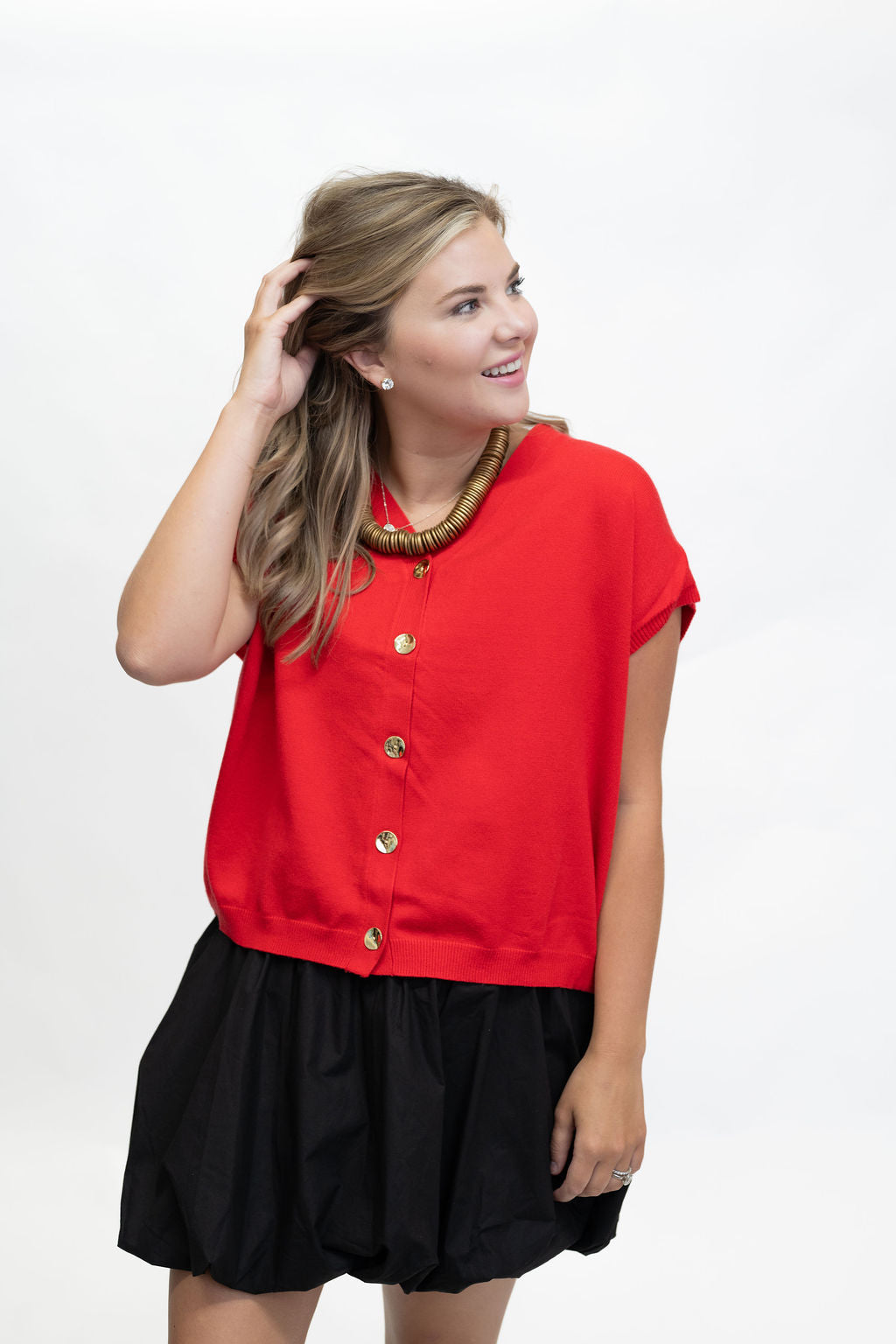 A woman modeling a bright red short-sleeve button-front top with gold buttons, paired with a black skirt, against a plain white studio background.