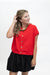 A woman modeling a bright red short-sleeve button-front top with gold buttons, paired with a black skirt, against a plain white studio background.