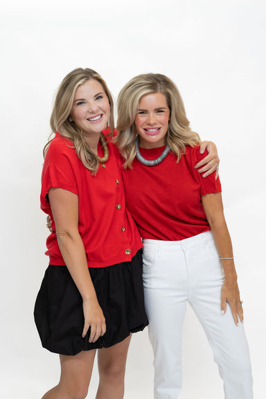 Two women posing together against a white studio background. The woman on the left wears a red short-sleeve button-up top with a black skirt, and the woman on the right wears a red short-sleeve top with white pants.
