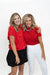 Two women posing together against a white studio background. The woman on the left wears a red short-sleeve button-up top with a black skirt, and the woman on the right wears a red short-sleeve top with white pants.