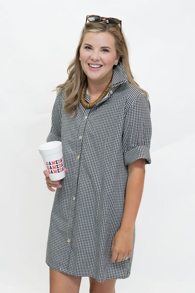 A woman wearing a black and white gingham short-sleeve button-up shirt dress, holding a white paper coffee cup with red and blue text, against a plain white background.
