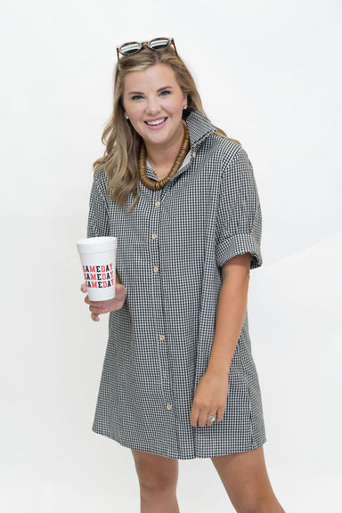 A woman modeling a short-sleeve black and white gingham button-down shirt dress, holding a white paper coffee cup with red and blue text that reads 'AMERICA MADE AMERICA'. She is standing against a plain white studio background.