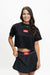 A woman modeling a black short-sleeve crop top and matching black skirt set against a plain white studio background. The set features a red rectangular chest patch with white text reading 'GO BARRIOS' and a matching waistband patch.