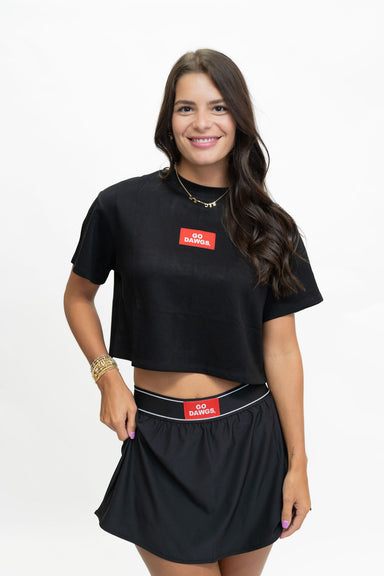 A woman modeling a black short-sleeve crop top and matching black skirt set against a plain white studio background. The set features a red rectangular chest patch with white text reading 'GO BARRIOS' and a matching waistband patch.