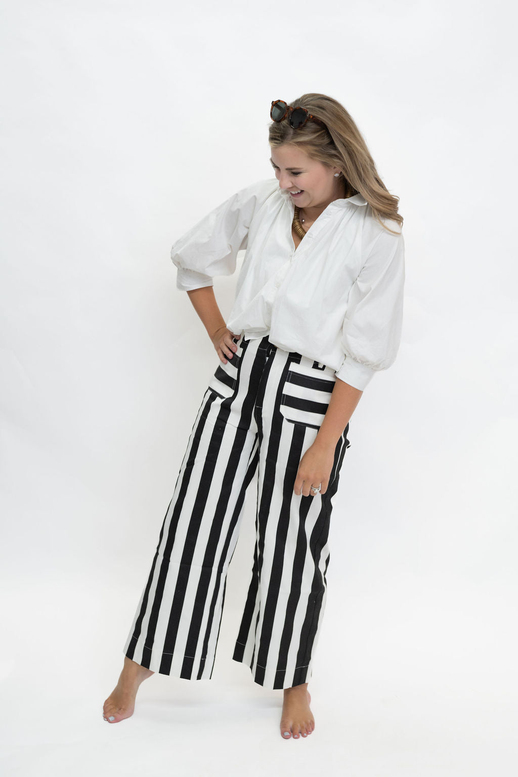 A woman posing in a white blouse and black and white vertically striped wide-leg pants against a plain white studio background.