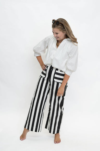 A woman posing in a white blouse and black and white vertically striped wide-leg pants against a plain white studio background.