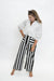 A woman posing in a white blouse and black and white vertically striped wide-leg pants against a plain white studio background.
