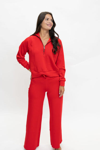A woman modeling a bright red two-piece tracksuit consisting of a long-sleeve half-zip sweatshirt and matching wide-leg pants, set against a plain white studio background.