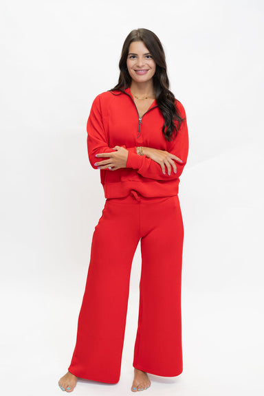 A woman modeling a bright red two-piece tracksuit with a half-zip long-sleeve top and matching wide-leg pants, standing against a plain white studio background.