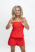 A woman modeling a bright red two-piece outfit consisting of a sleeveless crop top with thin shoulder straps and matching shorts, posed against a solid white studio background.