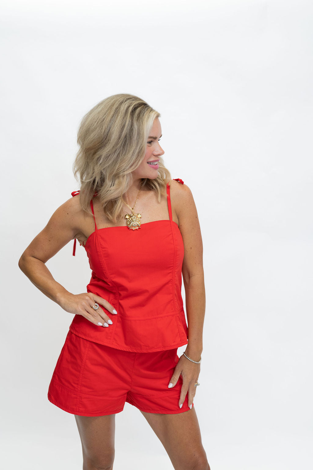 A woman modeling a bright red two-piece outfit consisting of a sleeveless crop top with thin shoulder ties and matching shorts, photographed against a plain white studio background.