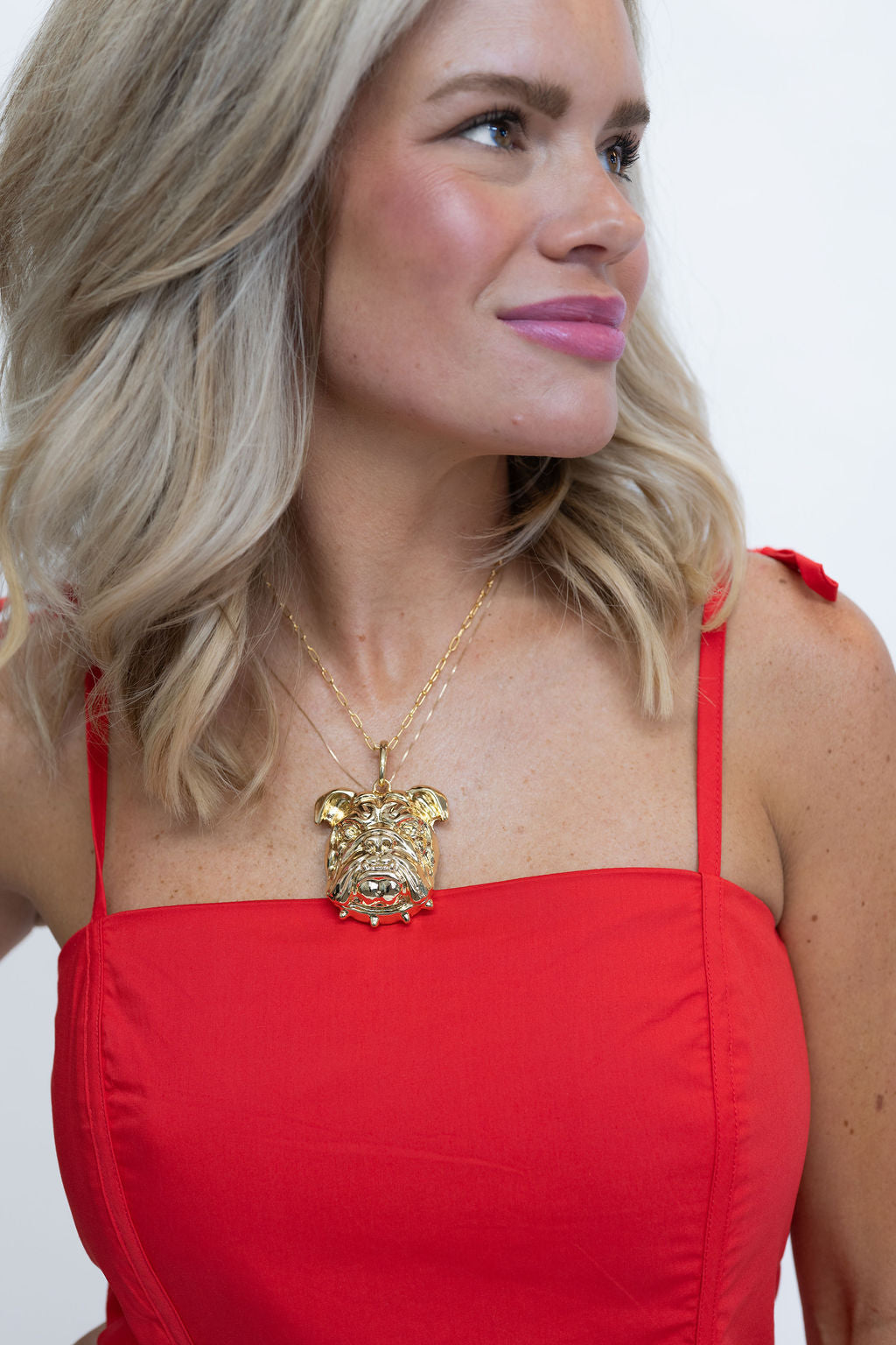 A blonde woman wearing a red strap dress and a gold bulldog pendant necklace, shown in a close-up profile against a plain white background.
