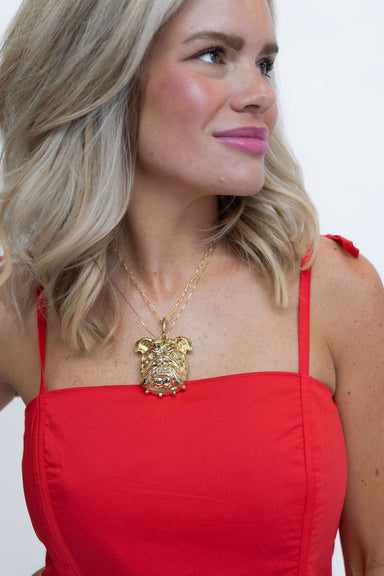 A blonde woman wearing a red strap dress and a gold bulldog pendant necklace, shown in a close-up profile against a plain white background.