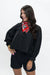 A woman modeling a black cropped jacket with balloon sleeves, paired with black shorts and a red patterned neck scarf, against a plain white studio background.
