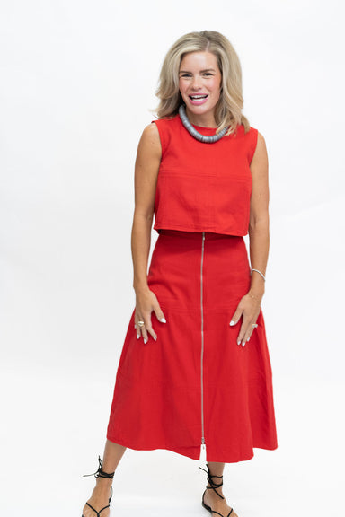 A woman modeling a bright red two-piece set consisting of a sleeveless cropped top and a matching midi skirt with a front zipper, styled with a chunky silver necklace and black strappy heels against a white studio background.