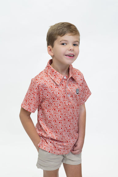 A young boy smiling and looking to the side, wearing a short-sleeve collared polo shirt with a red and white floral pattern, paired with beige shorts, against a plain white background.