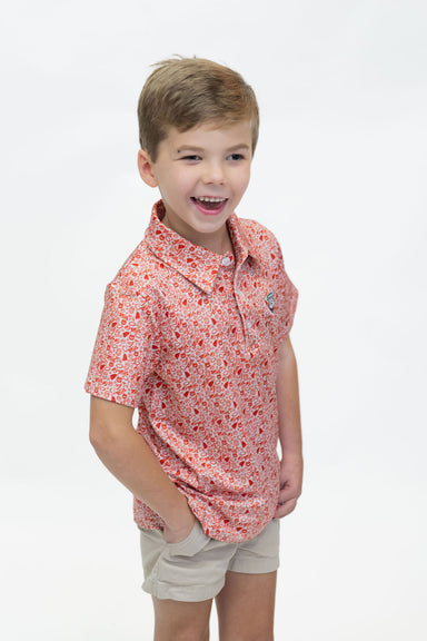 A young boy smiling and looking to the side, wearing a short-sleeve polo shirt with a red and orange floral pattern on a white background, paired with beige shorts, against a plain white studio background.