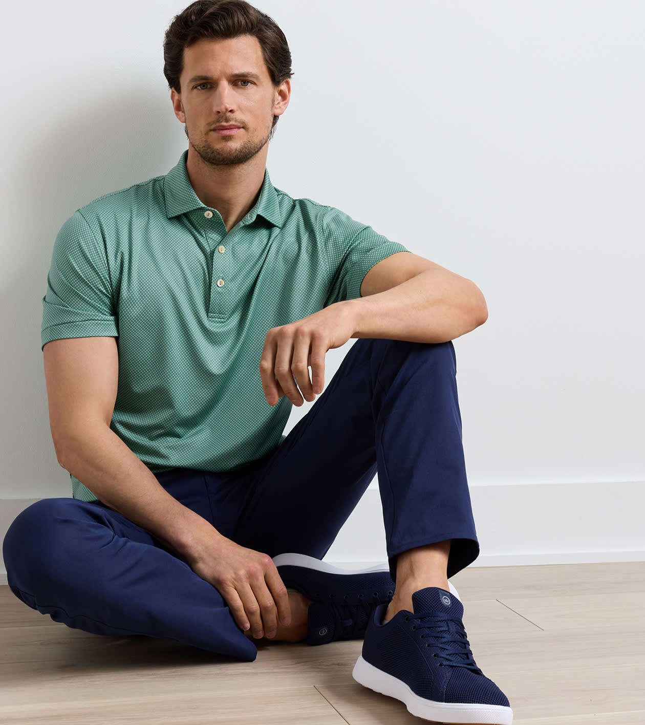 A man sitting cross-legged on a light wood floor against a white wall, wearing a sage green polo shirt, navy pants, and navy sneakers.