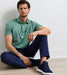 A man sitting cross-legged on a light wood floor against a white wall, wearing a sage green polo shirt, navy pants, and navy sneakers.