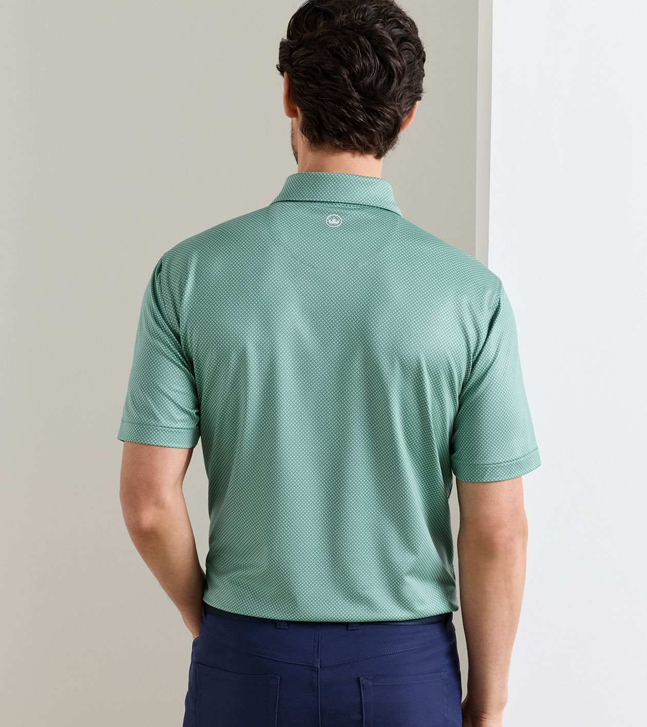 A man viewed from behind wearing a sage green short-sleeve polo shirt and navy blue pants, standing against a neutral beige wall.