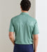 A man viewed from behind wearing a sage green short-sleeve polo shirt and navy blue pants, standing against a neutral beige wall.