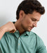 A close-up of a man wearing a textured sage green short-sleeve polo shirt, looking down and adjusting the collar button with his hand.