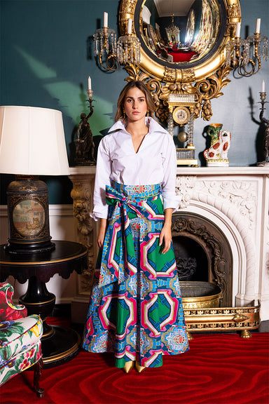 Woman in a colorful dress standing in an ornate room with a fireplace and decorative mirror.