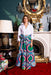 Woman in a colorful dress standing in an ornate room with a fireplace and decorative mirror.