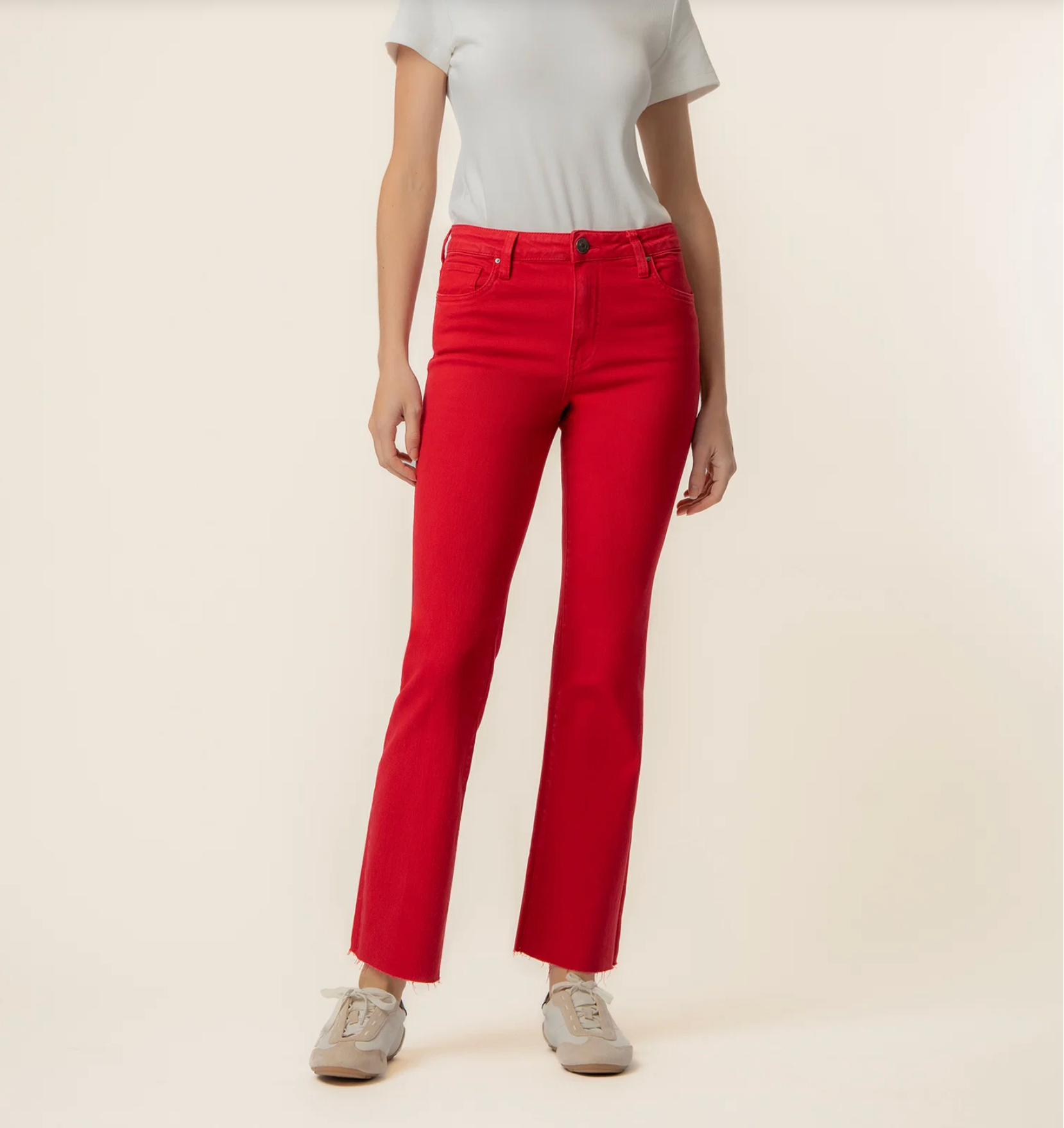 A model wearing bright red ankle-length jeans, a light gray t-shirt, and gray sneakers, standing against a solid light beige background.