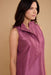 A woman wearing a sleeveless, high-neck satin-like top in a dusty mauve or plum purple color, shown from the chest up against a solid beige studio background.