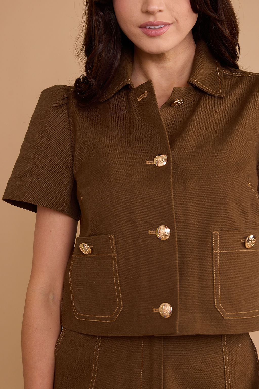 A close-up of a model wearing a brown short-sleeve button-front jacket with gold-tone buttons and contrast stitching, paired with a matching brown skirt or pants against a warm beige background.