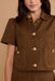 A close-up of a model wearing a brown short-sleeve button-front jacket with gold-tone buttons and contrast stitching, paired with a matching brown skirt or pants against a warm beige background.