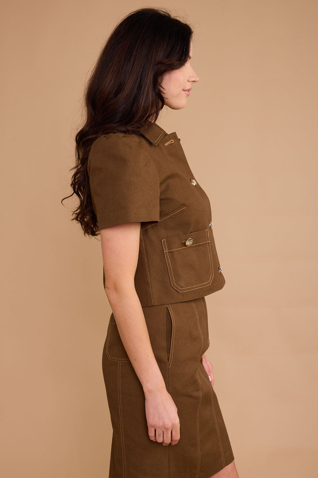 A woman shown in side profile wearing a brown short-sleeve cropped vest and matching brown skirt against a plain beige studio background.